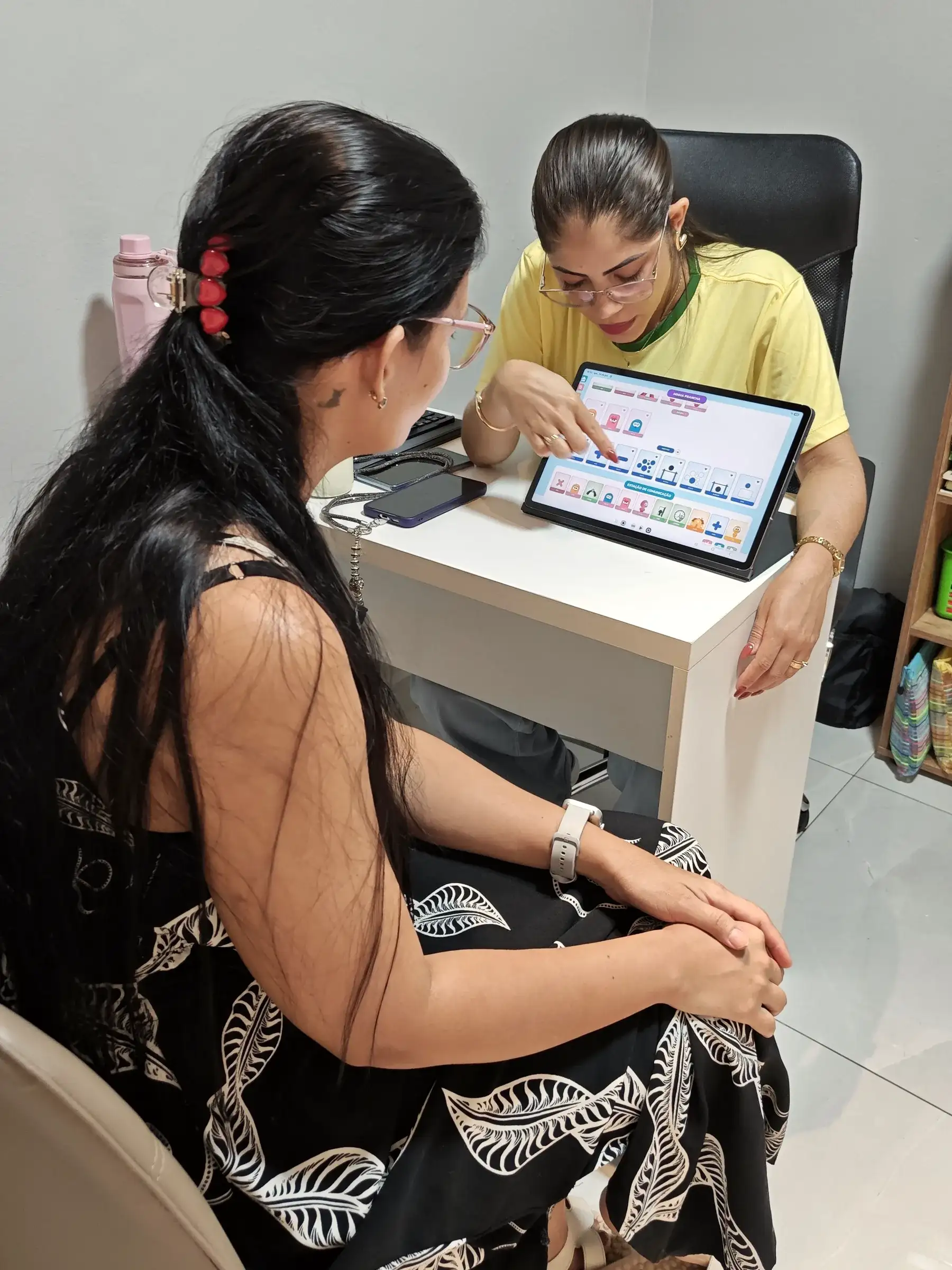 Profissional da Progride mostra a uma mãe uma prancha de comunicação no tablet, em uma sala de atendimento