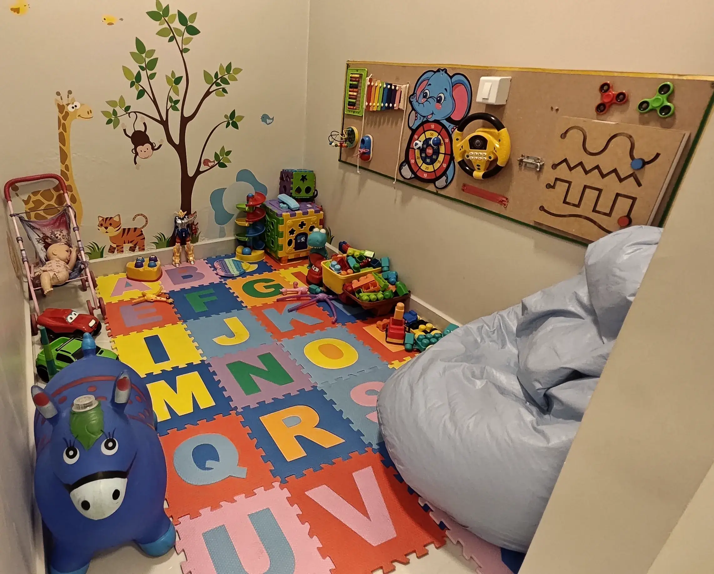 Sala lúdica com tapete de letras, brinquedos e painel sensorial na parede